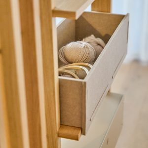 Nahaufnahme eines WoodUpp-Wandregals: schlanke Eichenholz-Konstruktion mit herausziehbarer, grauer Filzschublade, gefüllt mit beigen Wollknäueln und Holzstickrahmen. Die weiche Filzbox schützt die Wolle, sichtbare Holzleisten und Zapfen betonen die modulare, minimalistische, handgefertigte Aufbewahrungslösung von WoodUpp, mit warmem Holzton, ideal für Handarbeiten.