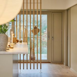 WoodUpp Raumteiler aus schlanken, vertikalen Eichenlatten mit dezenten Metallfüßen und quer verlaufenden Holzleisten, an denen Holz‑Haken und Netztasche hängen. Der helle, minimalistische Raumteiler trennt Küche und Flur, lässt Licht durch, bietet dezente Ablage- und Aufhängemöglichkeiten sowie leichten Blickschutz mit natürlichem Eichenfinish.