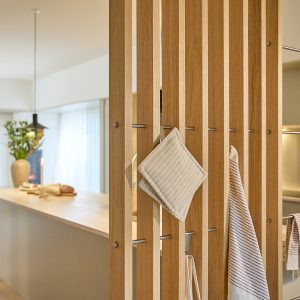 WoodUpp Holzlamellen-Raumteiler aus Eichenholz mit feiner Maserung und warmem, natürlichem Ton, ausgestattet mit schlanken Edelstahlstangen als Haken. An ihnen hängen ein gestreifter Topflappen, ein Geschirrtuch und eine Schürze. Im Hintergrund erkennbar: helle Küche mit großer Arbeitsinsel, Pendelleuchte, Vase sowie Brot.