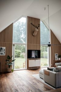 Helles Wohnzimmer mit hoher Giebelfront, WoodUpp-Lattenwand aus schlanken, vertikalen Holzlamellen und großem Fenster, montiertem Fernseher sowie Hirschgeweih als Wanddekoration. Auf dem Boden WoodUpp-Holzparkett mit warmer, gelebter Maserung, dazu Sideboard, Pflanze und graues Sofa — helles Tageslicht und Blick in den Garten.