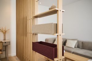 WoodUpp Raumteiler-Regal aus hellem, naturbelassenem Eichenholz mit schlanken Lamellen, modularem Holzrahmen und sichtbaren Metallbolzen; daran zwei WoodUpp Filzboxen, offene rechteckige, tiefe Aufbewahrungsboxen in Beige und Bordeauxrot sowie ein kleines Regalbrett mit Dekoobjekten. Im Hintergrund Sofa, Beistelltisch und Arbeitsplatz, minimalistisches skandinavisches Design.