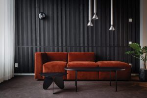 Modernes Wohnzimmer mit rostrotem Samtsofa vor einer WoodUpp Lamellenwand aus schwarzen, vertikalen Holzlamellen. Die WoodUpp Wandpaneele zeigen feine Schattenfugen und tiefe Struktur. Vor dem Sofa stehen niedrige schwarze Couchtische, drei silberne Hängeleuchten und ein dunkler Teppich runden die elegante Szenerie ab.