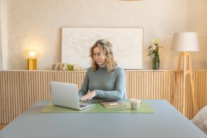 Eine Frau arbeitet an einem Laptop auf einer grünen WoodUpp-Schreibtischunterlage; daneben liegen ein Notizbuch und eine Tasse auf einem großen hellgrauen Tisch. Im Hintergrund ein helles Sideboard mit Holzlamellen, eine Vase mit Blumen sowie zwei WoodUpp-Holzlampen (Tisch- und Stehleuchte) im warmen, skandinavischen Stil.