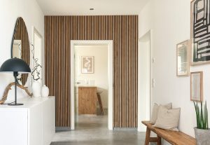 Heller Flur mit WoodUpp-Lamellenwand: vertikale Holzlamellen als warme Wandverkleidung um eine Türöffnung, kombiniert mit weißer Kommode, rundem Wandspiegel, schwarzer Tischlampe und Holzbänkchen. WoodUpp-Lamellenpaneele wirken als dezente, moderne Akzentwand. Die schmalen, gleichmäßig geführten Lamellen zeigen natürliche Holzmaserung und schaffen Tiefe im Raum.