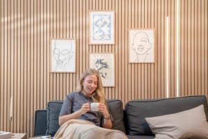 Frau sitzt auf dunkelgrauem Sofa, mit Decke und einer Tasse, vor einer WoodUpp Lamellen-Holzwand mit schlanken, vertikalen Lamellen. An der Wand sind vier gerahmte Bilder und zwei integrierte, schlanke WoodUpp LED-Lichtleisten, die warmes Licht im natürlichen Farbton spenden.