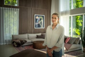 Eine lächelnde Frau steht in einem lichtdurchfluteten Wohnzimmer vor einer großflächigen WoodUpp-Lamellenwand aus hellem Holz; im Vordergrund ein runder, gerippter WoodUpp-Holzcouchtisch. Beiges Ecksofa mit Kissen und Decke, gemusterter Teppich, große Fenster mit Gardinen und ein Kaktus komplettieren die moderne, warme Einrichtung.
