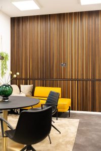 Modernes Lounge-Interieur mit markanter WoodUpp-Lamellenwand: schmale, vertikale, warmholzfarbene Lamellenpaneele von WoodUpp als Wandverkleidung mit schwarzen Fugen und horizontaler Zierleiste. Im Vordergrund gelbes Sofa mit Kissen, zwei schwarze Stühle, runder Esstisch, beiger Teppich, grüne Glasvase, Pflanze; sichtbare Steckdosen und elegantes Metallgestell runden das minimalistische Design ab.