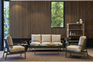 WoodUpp: Ruhiges Lounge-Interieur mit markanter WoodUpp-Wandverkleidung aus vertikalen, dunkel-hell akzentuierten Holzlamellen, einem schlanken WoodUpp-Regal aus hellem Holz, einem dreisitzigen Sofa und zwei Sesseln mit olivegrünen Metallrahmen und sandfarbenen Polsterkissen, bequemen, voluminösen Polstern, grünem Pflanzenakzent, goldenen Trinkgläsern auf Tablett, großem Fenster mit üppigem Grünblick im Hintergrund.