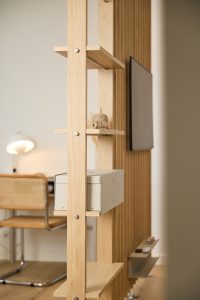 WoodUpp-Raumteiler aus hellem Eichenholz mit vertikalen Lamellen, modularen Regalböden, sichtbaren Metallverschraubungen und offener Ablage, dezenter Beleuchtung sowie einer hellgrauen Metall-Schublade von WoodUpp; an der Rückseite hängt ein Flachbildschirm, auf einem Regal liegt eine Garnrolle, im Hintergrund Schreibtisch mit geflochtenem Stuhl.
