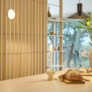 WoodUpp Lamellenwand aus hellen Eichenleisten mit integrierten offenen Regalböden und kleinen Metallhaken, im Bild als Raumteiler neben einer großen Fensterfront. Auf den Regalböden kleine Keramikschalen und Dekostücke, dazu eine runde Wandlampe; im Vordergrund Kerze und Brot, warme, minimalistische Wohnatmosphäre.