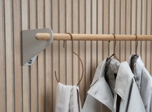 WoodUpp Lamellenwand aus hellem Eichenholz mit schmalen, senkrechten Lamellen und sichtbarer Holzmaserung, kombiniert mit einer WoodUpp Garderobenstange: runde, geölte Holzstange auf dezenter sandgrauer Metallhalterung. An der Stange hängen Leder-Schlaufenhaken, ein leichter Schal und zwei helle Mäntel, elegant in warmen Naturtönen.