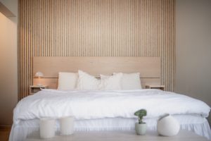 Schlafzimmer mit WoodUpp-Produkten: minimalistischem Design, helle WoodUpp-Eichen-Lamellen-Wandverkleidung mit feinen vertikalen, raumhohen Lamellen von Boden bis Decke, passendes WoodUpp-Kopfteil in natürlichem Holzton und zwei schlichte WoodUpp-Holz-Nachttische. Weiß bezogenes Bett mit weicher Leinenbettwäsche, Kissen, links kleine Tischlampe, vorn Kerzen, Vase und kleine Zimmerpflanze.