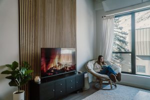 Moderner Wohnraum mit WoodUpp Holzlamellen-Akustikpaneel an der Wand hinter einem Fernseher; die hellbraunen vertikalen Lamellen schaffen Struktur. Auf der dunklen TV-Kommode flackert ein Kaminfilm, daneben Pflanze und Korb-Sessel mit Kissen, große Fenster bringen viel Tageslicht. Eine Frau liest entspannt und bequem.