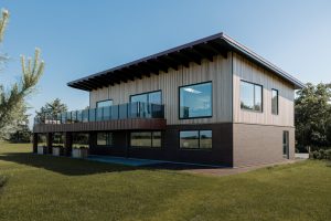 Modernes zweistöckiges Haus mit WoodUpp-Holzlamellen an der oberen Fassade, klaren großen Fenstern und einem überdachten Balkon. Zu sehen sind WoodUpp senkrechte, fein profilierte Fassadenlatten in heller Holzoptik sowie Holzverkleidung am Balkonrand, kombiniert mit Glasbrüstung, Backstein-Sockel und grünem Rasen unter blauem Himmel.