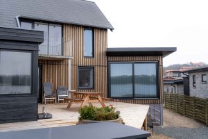 Modernes Haus mit WoodUpp-Fassadenlamellen: vertikale Holzlatten an der Fassade, große schwarze Fenster und eine WoodUpp-Terrasse aus Dielen. Auf dem Holzdeck steht ein Picknicktisch, zwei Klappstühle und ein Pflanzenkübel; im Fenster spiegelt sich ein See. Ein kleiner Balkon mit Metallgeländer oberhalb.